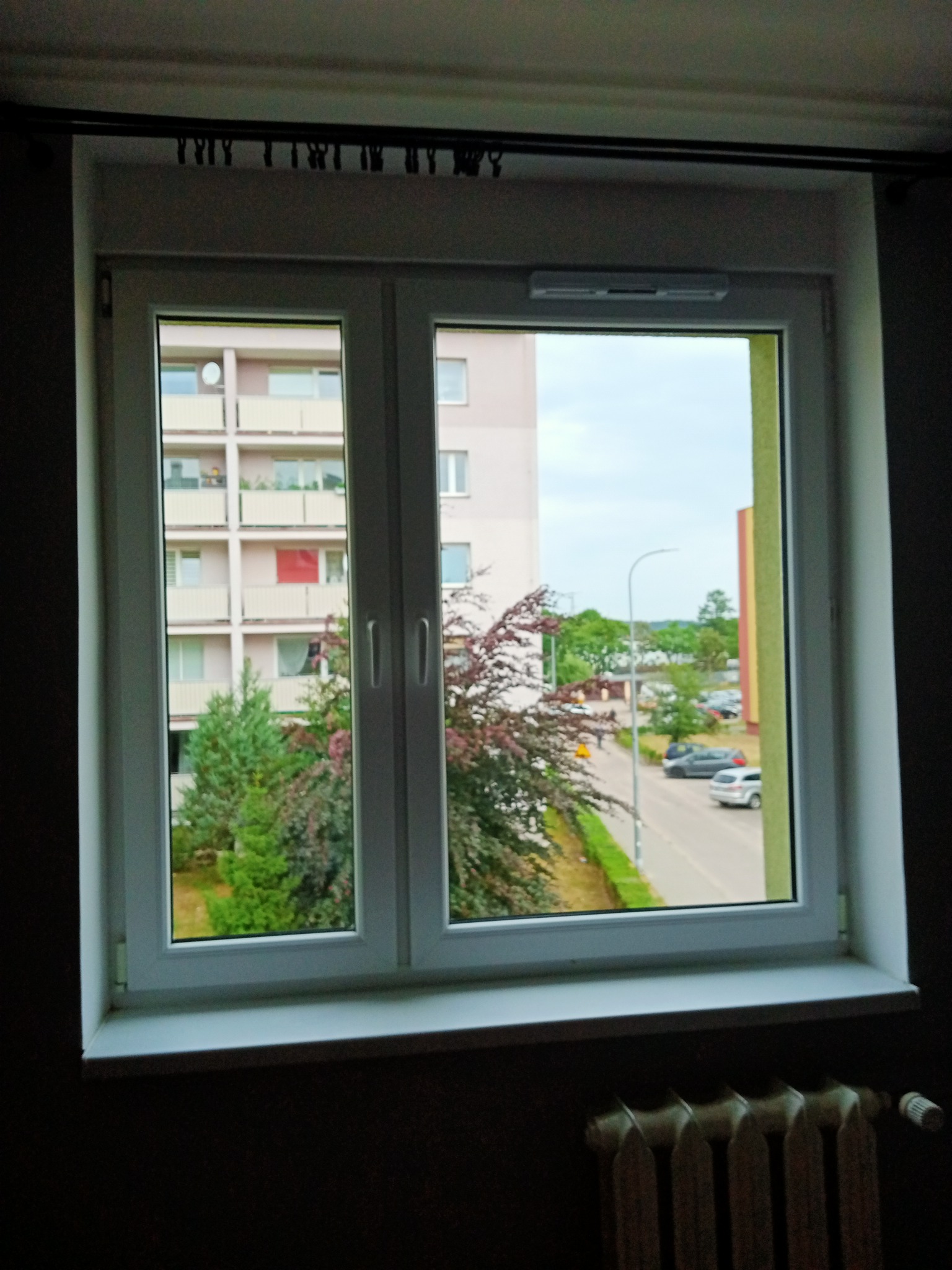 Widok z okna mieszkania na blok z balkonami i ulicę z samochodami w pochmurny dzień.