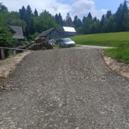 Droga gruntowa pokryta szarym tłuczniem, w tle wiejskie zabudowania, sterta drewna i zaparkowany srebrny samochód osobowy.
