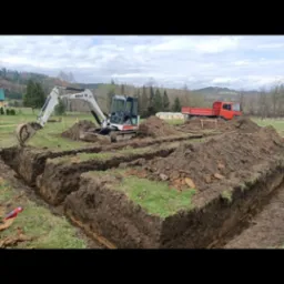 Biała minikoparka Bobcat na placu budowy, wykonująca wykopy pod fundamenty, z czerwoną ciężar&oacute;wką w tle i zielonymi wzg&oacute;rzami na horyzoncie.