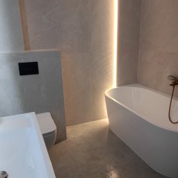 Sap-Tom Instalacje Elektryczne, Wykończenia - Nowoczesna łazienka z wanną, umywalką i toaletą w odcieniach szarości, podświetlona pionowym paskiem LED. Minimalistyczny design z marmurkowymi płytkami.