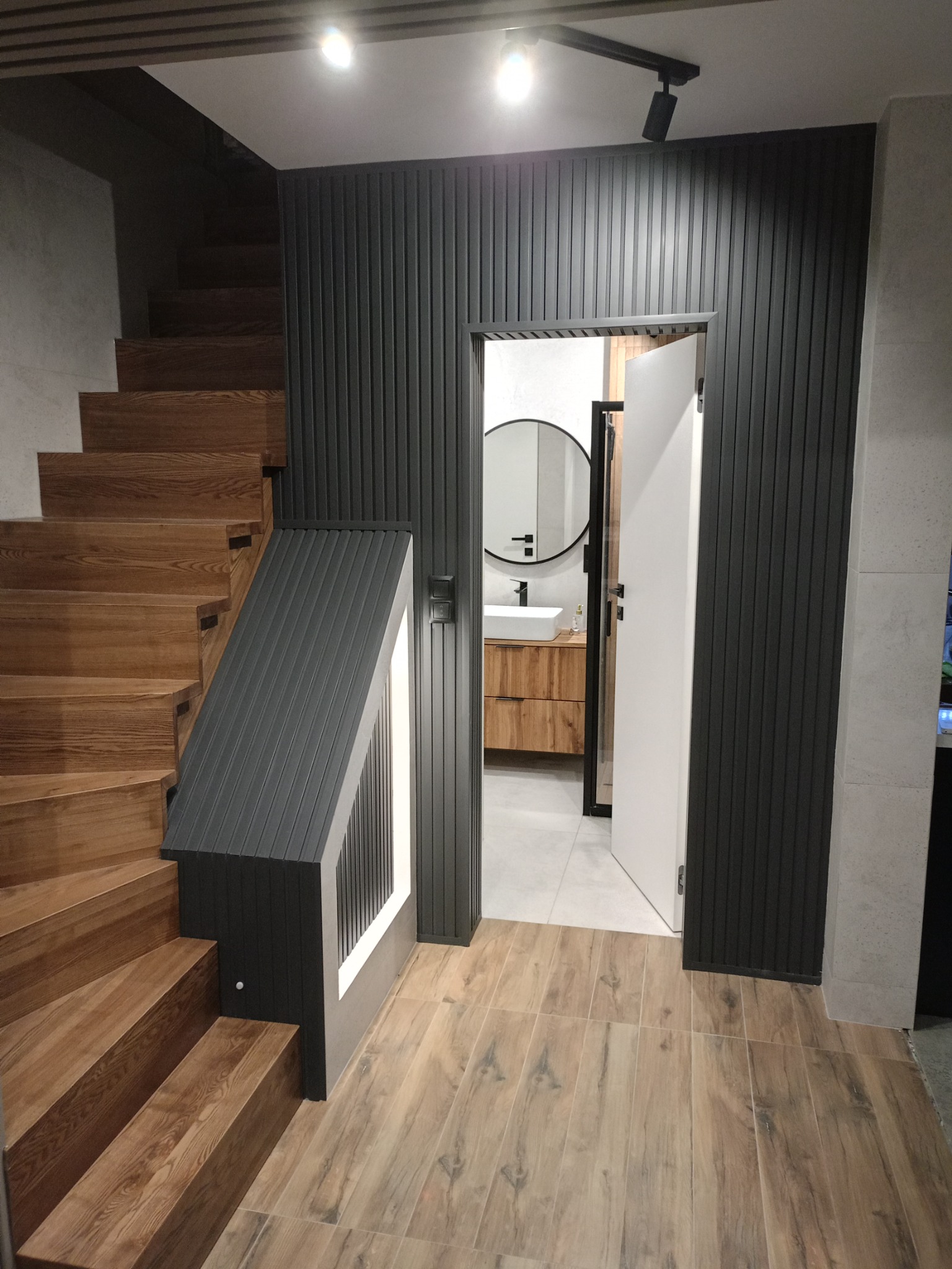 Drewniane schody z podświetleniem, ciemna ściana z paneli pionowych, otwarte drzwi do łazienki z okrągłym lustrem i drewnianą szafką pod umywalką.