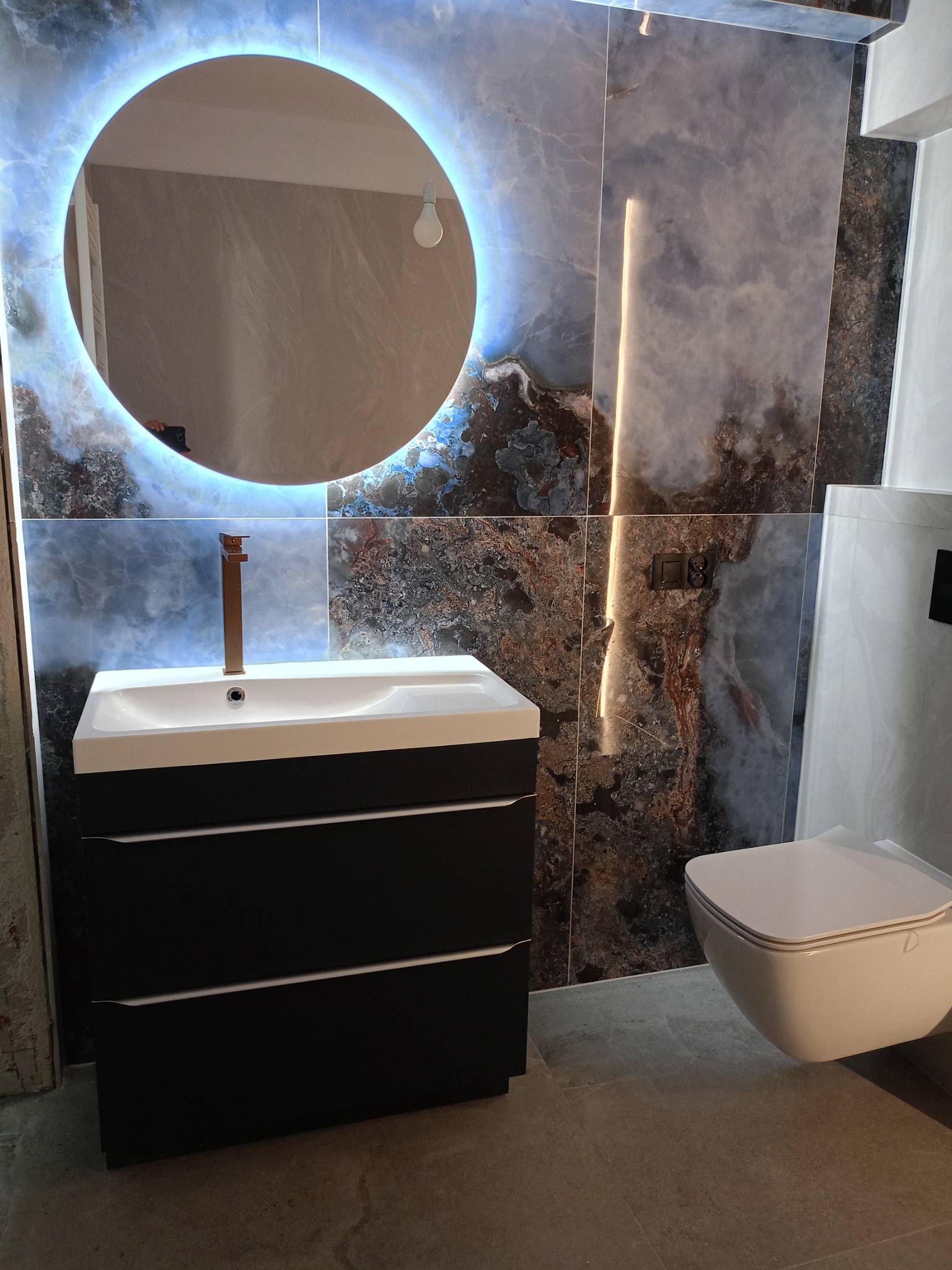 Nowoczesna łazienka: czarna szafka z umywalką, okrągłe lustro z podświetleniem LED na tle marmurowych płytek. Minimalistyczna toaleta podwieszana.