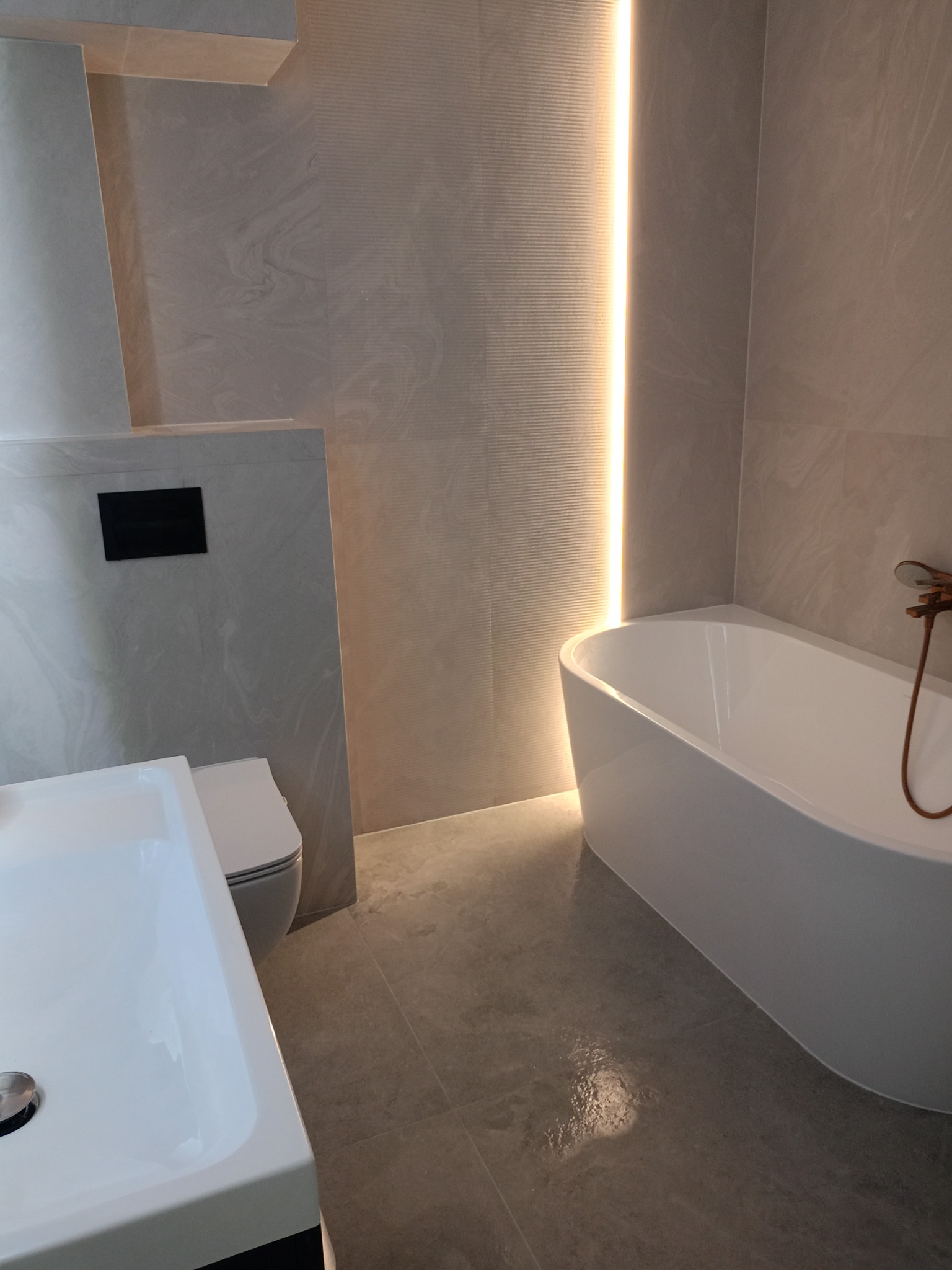 Nowoczesna łazienka z wanną, umywalką i toaletą w odcieniach szarości, podświetlona pionowym paskiem LED. Minimalistyczny design z marmurkowymi płytkami.