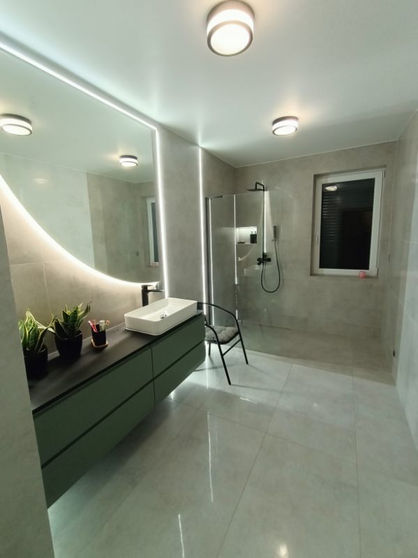 Nowoczesna łazienka z podświetlanym lustrem, zieloną szafką pod umywalką i kabiną prysznicową walk-in, wykończona w szarościach z akcentami roślinnymi.