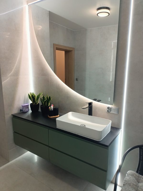 Nowoczesna łazienka: zielona szafka pod umywalką, biała prostokątna umywalka, czarna bateria, lustro z podświetleniem LED, rośliny doniczkowe.