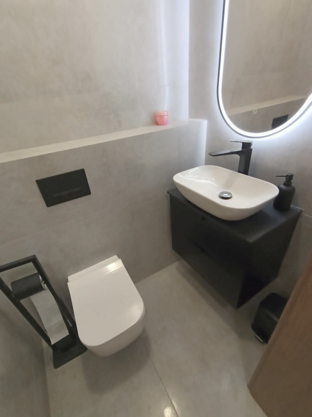 Nowoczesna łazienka w szarościach: podwieszana toaleta, umywalka na szafce i podświetlane lustro owalne. Minimalistyczny design z akcentem w postaci różowego akcentu.