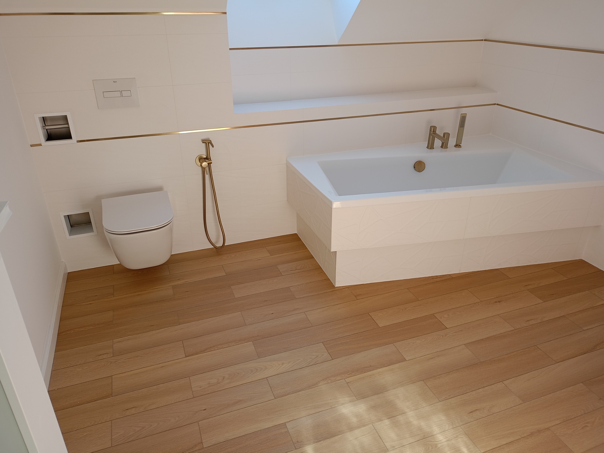 Biała łazienka z drewnianą podłogą, złotymi akcentami i nowoczesną armaturą. Widoczna wanna, toaleta i bidetka. Uwagę przykuwają detale w kolorze złota.