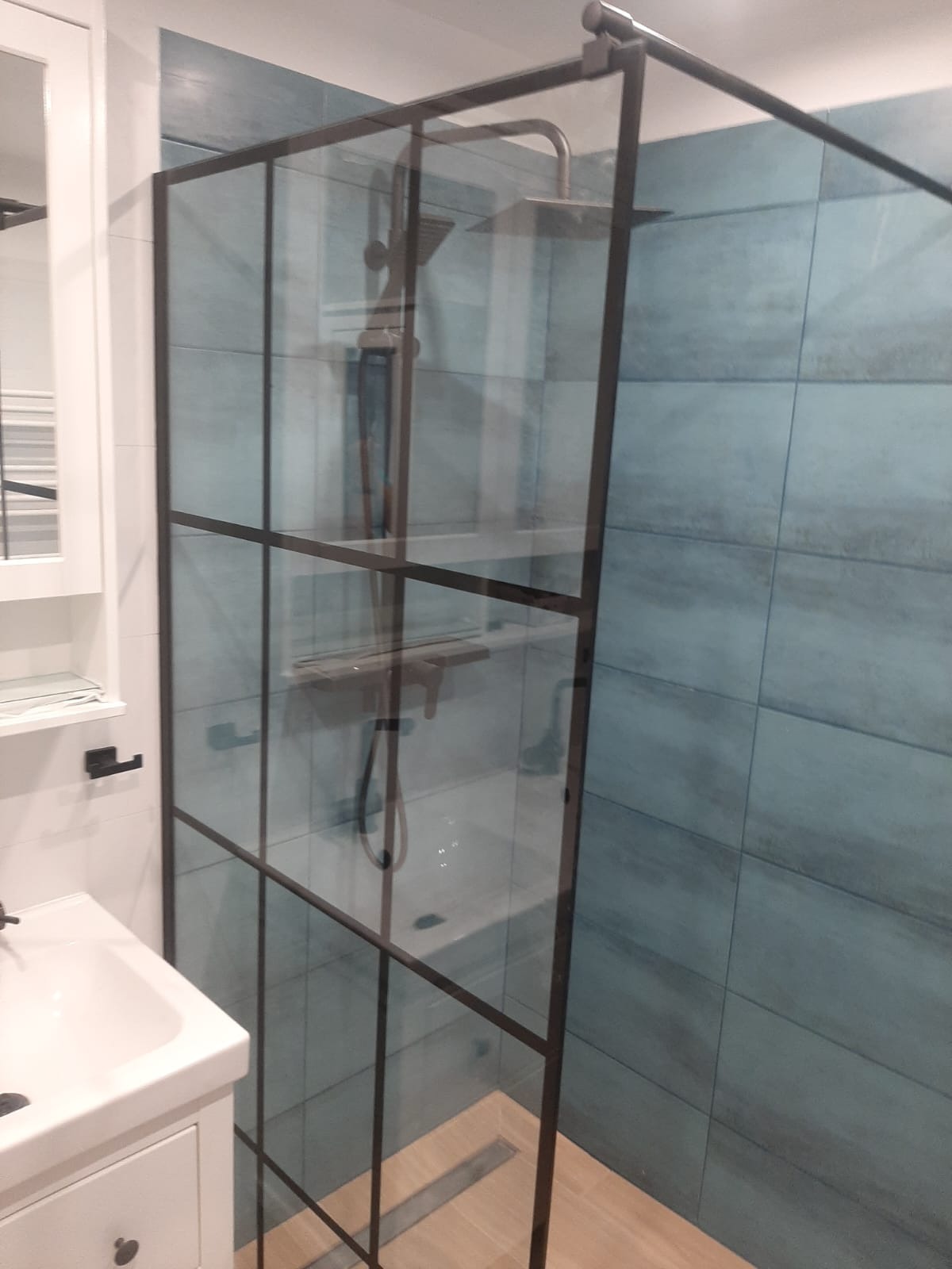 Nowoczesna kabina prysznicowa z czarnymi profilami na tle ściany wyłożonej turkusowymi płytkami, obok biała umywalka z szafką.