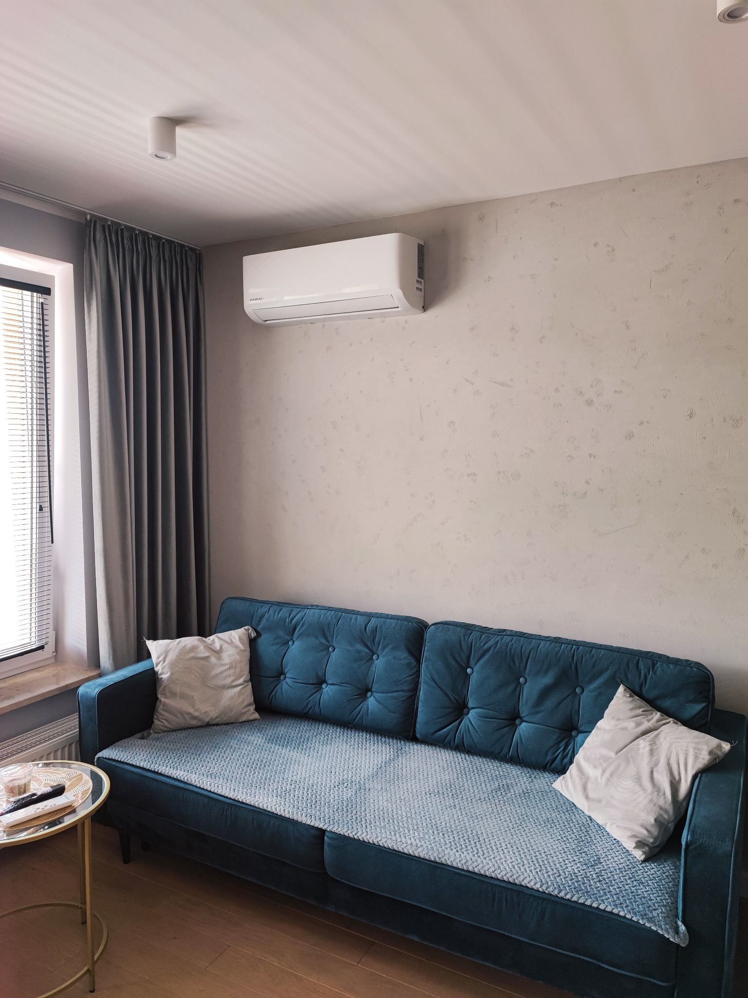 Biały klimatyzator ścienny zamontowany nad turkusową sofą w pokoju z beżowymi ścianami, obok okna z żaluzjami i szarymi zasłonami.