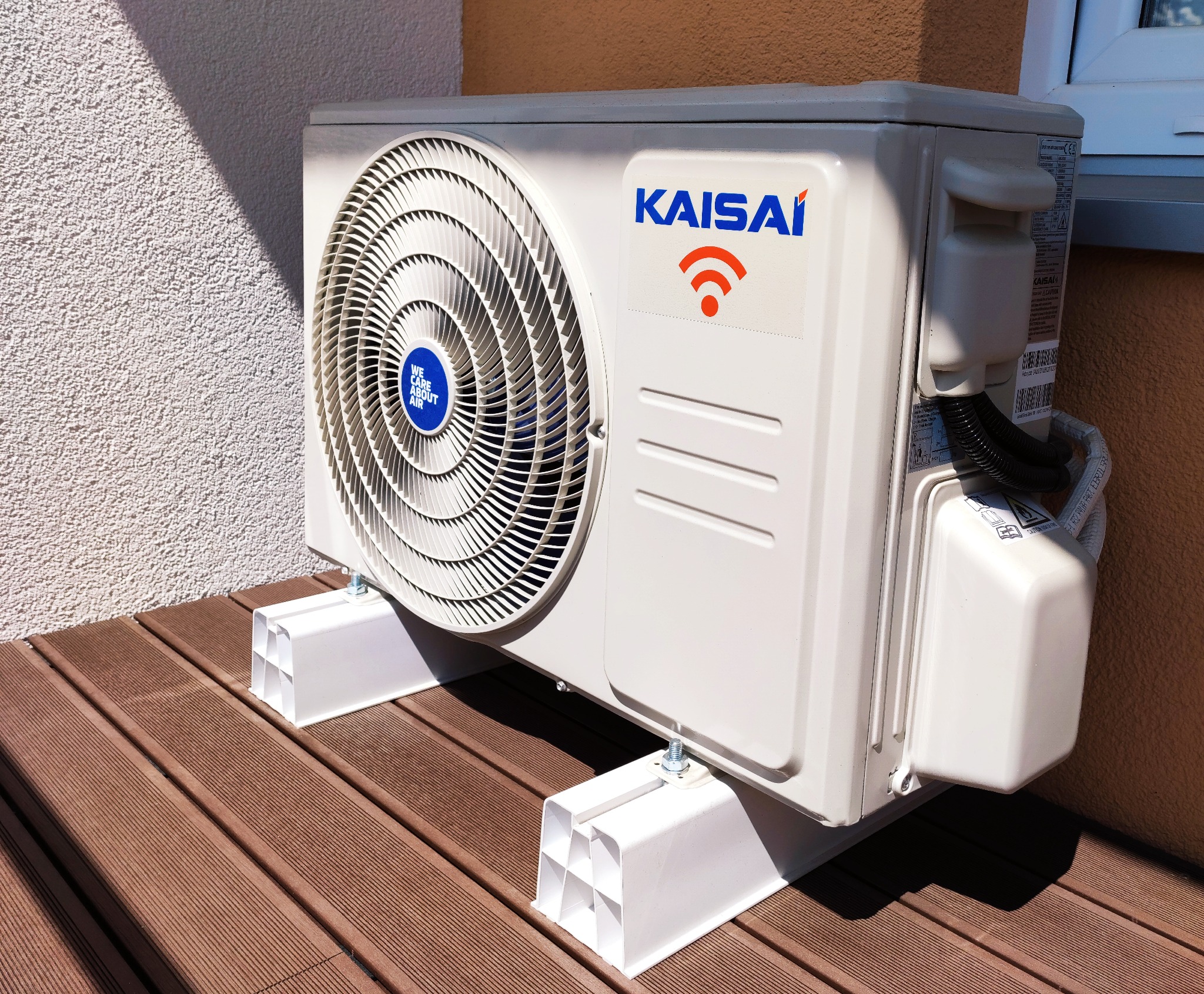 Zewnętrzna jednostka klimatyzacji Kaisai zamontowana na białych wspornikach na brązowym tarasie kompozytowym, z widocznym logiem WiFi i napisem 'We Care About Air'.