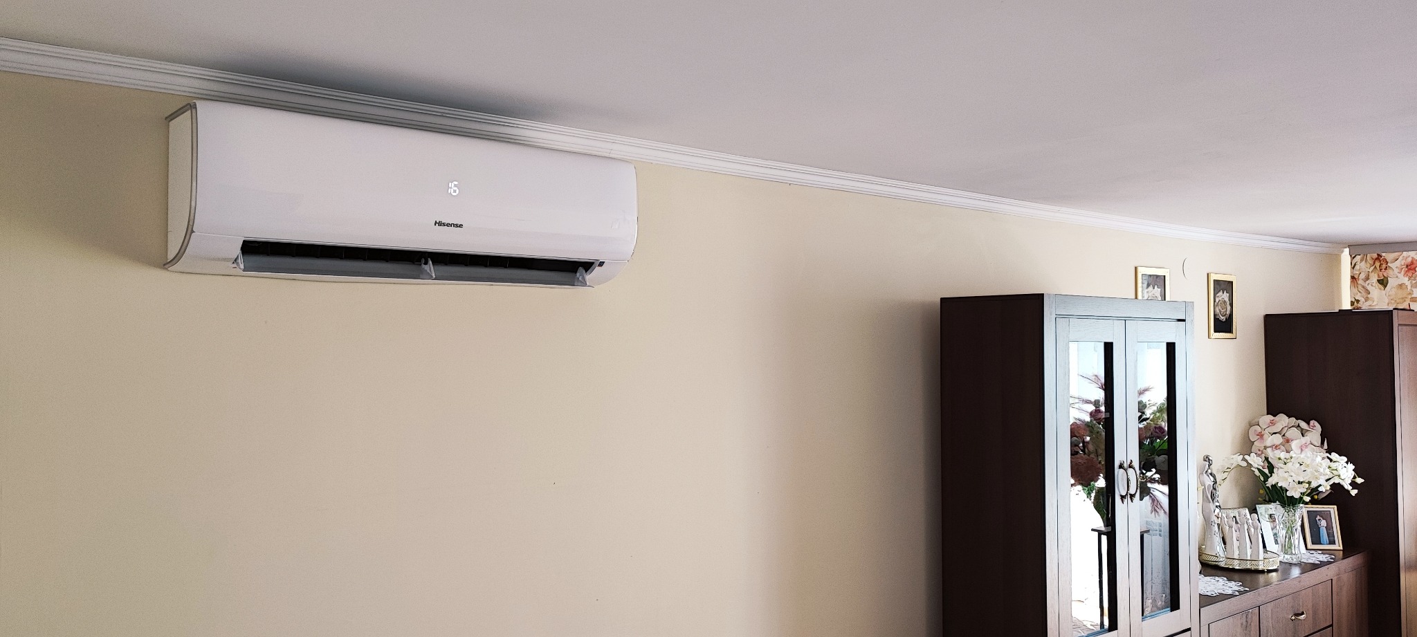 Biały klimatyzator marki Hisense zamontowany na ścianie w pokoju z beżową ścianą, ozdobnym gzymsem sufitowym i meblami w tle.