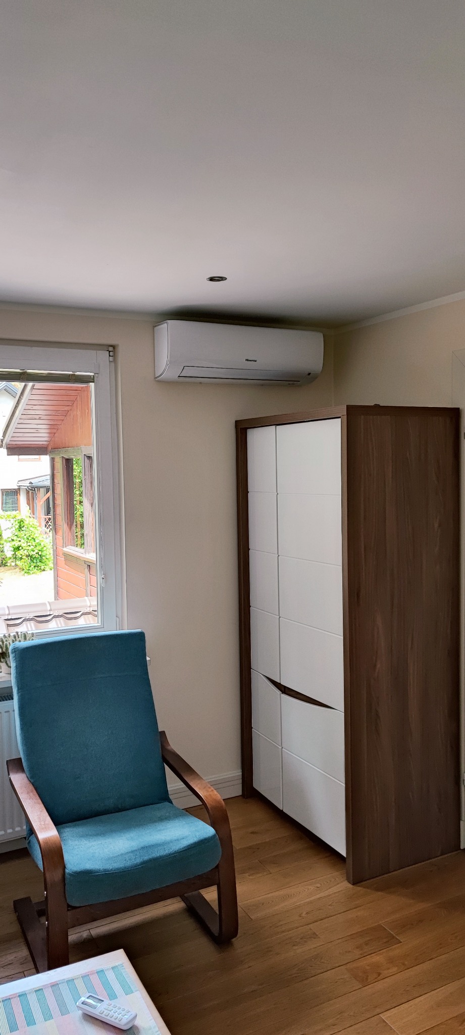 Nowoczesny klimatyzator Hisense zamontowany na ścianie w pokoju, obok okna i szafy, widok na zewnątrz przez okno.