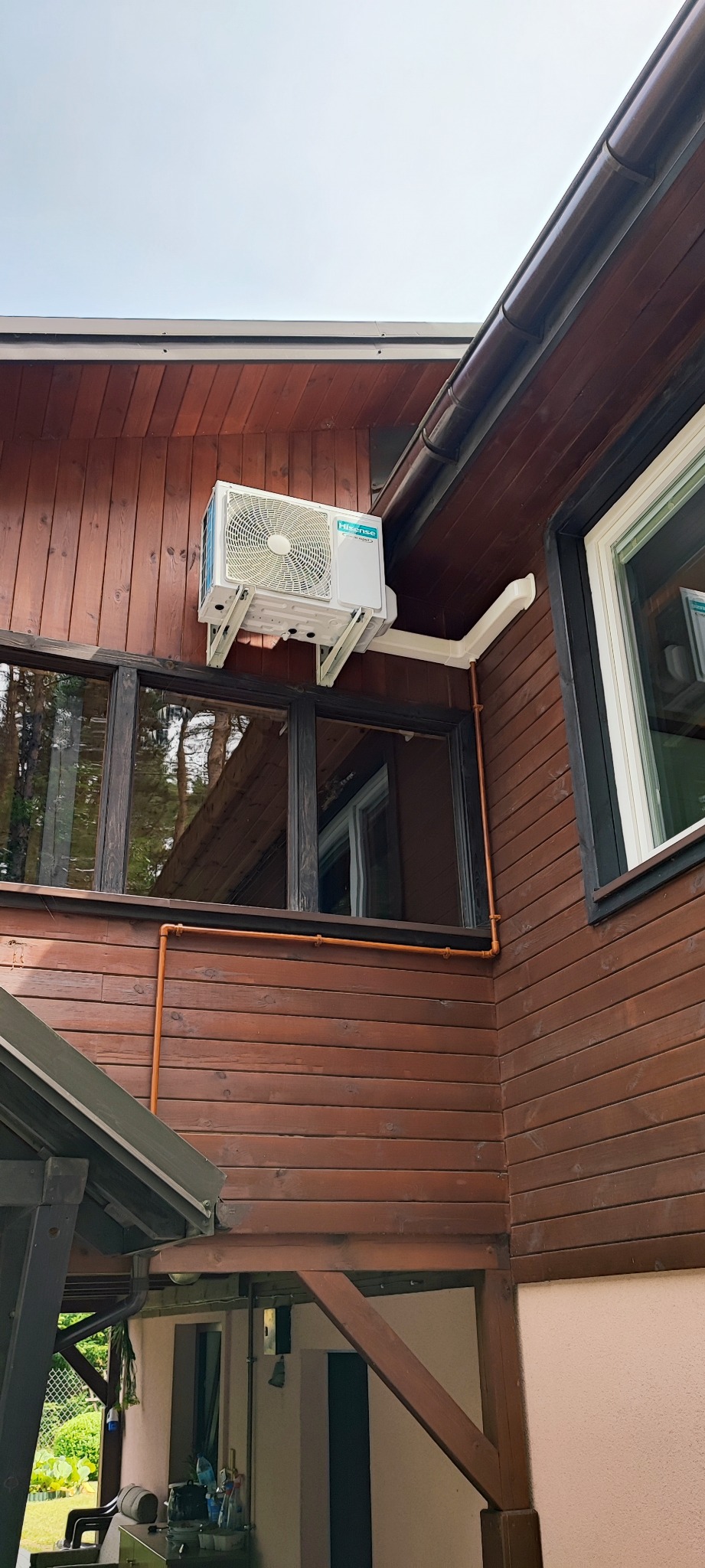 Zewnętrzna jednostka klimatyzacji Hisense zamontowana na drewnianej ścianie domu, widoczne rury odprowadzające skropliny i okno z czarną ramą.