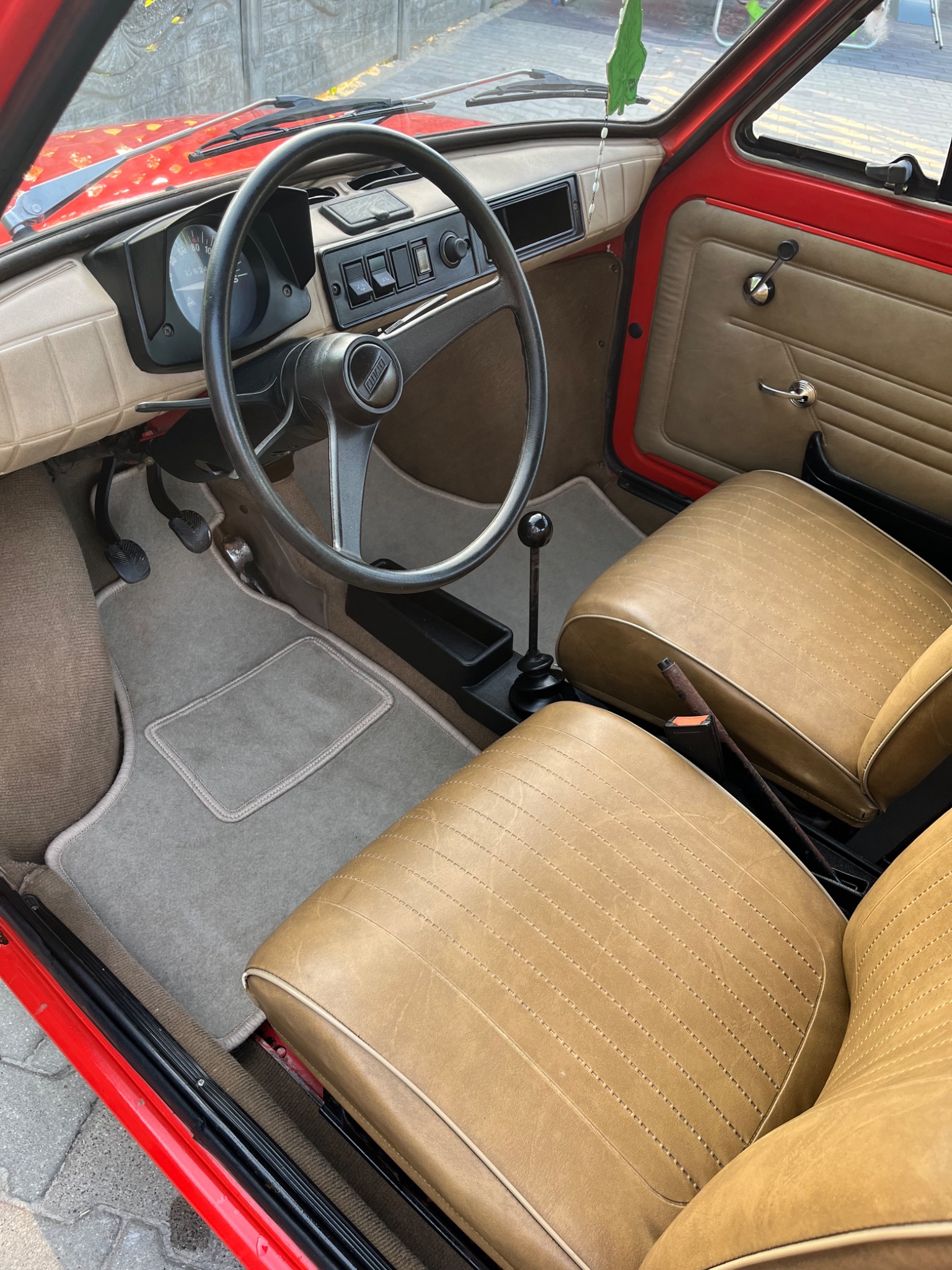 Wnętrze czerwonego Fiata 126p z beżową tapicerką skórzaną, widoczne fotele, deska rozdzielcza, kierownica i dywaniki