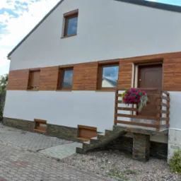 Dom z białą elewacją, drewnianą okładziną i kamienną podmurówką, z drewnianą balustradą i kwiatami przy wejściu.