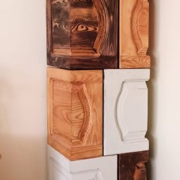 Pawel Manka - Nietypowa kolumna z drewnianych, kwadratowych modułów z dekoracyjnymi frezami, w kolorach naturalnego drewna i bieli, ustawiona na niskim stoliku z giętymi nogami.