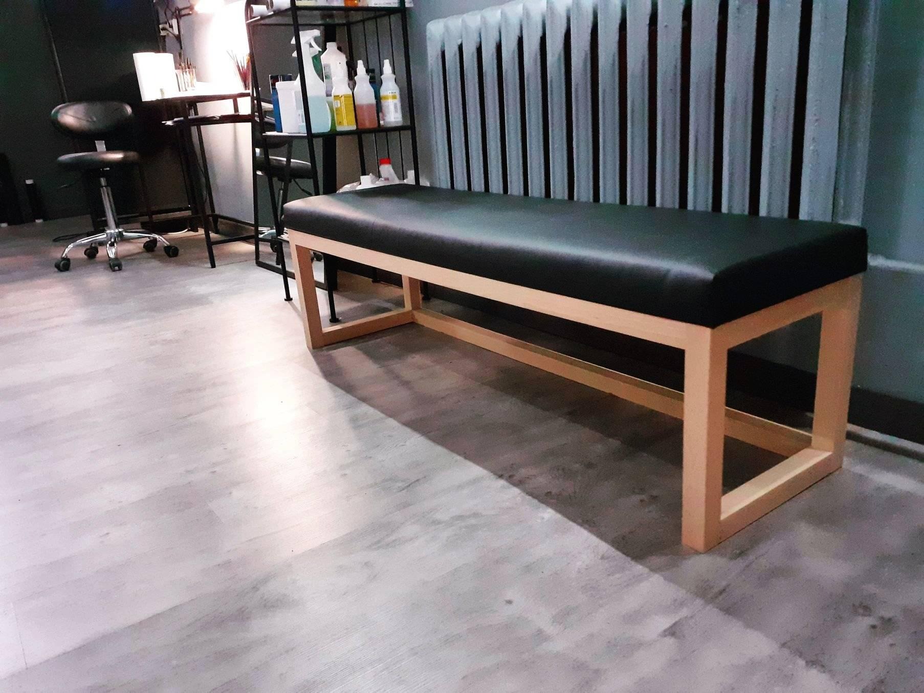 Prosty, drewniany stół z czarnym, tapicerowanym siedziskiem w pomieszczeniu z szarymi ścianami i podłogą, obok metalowy regał i krzesło na kółkach.