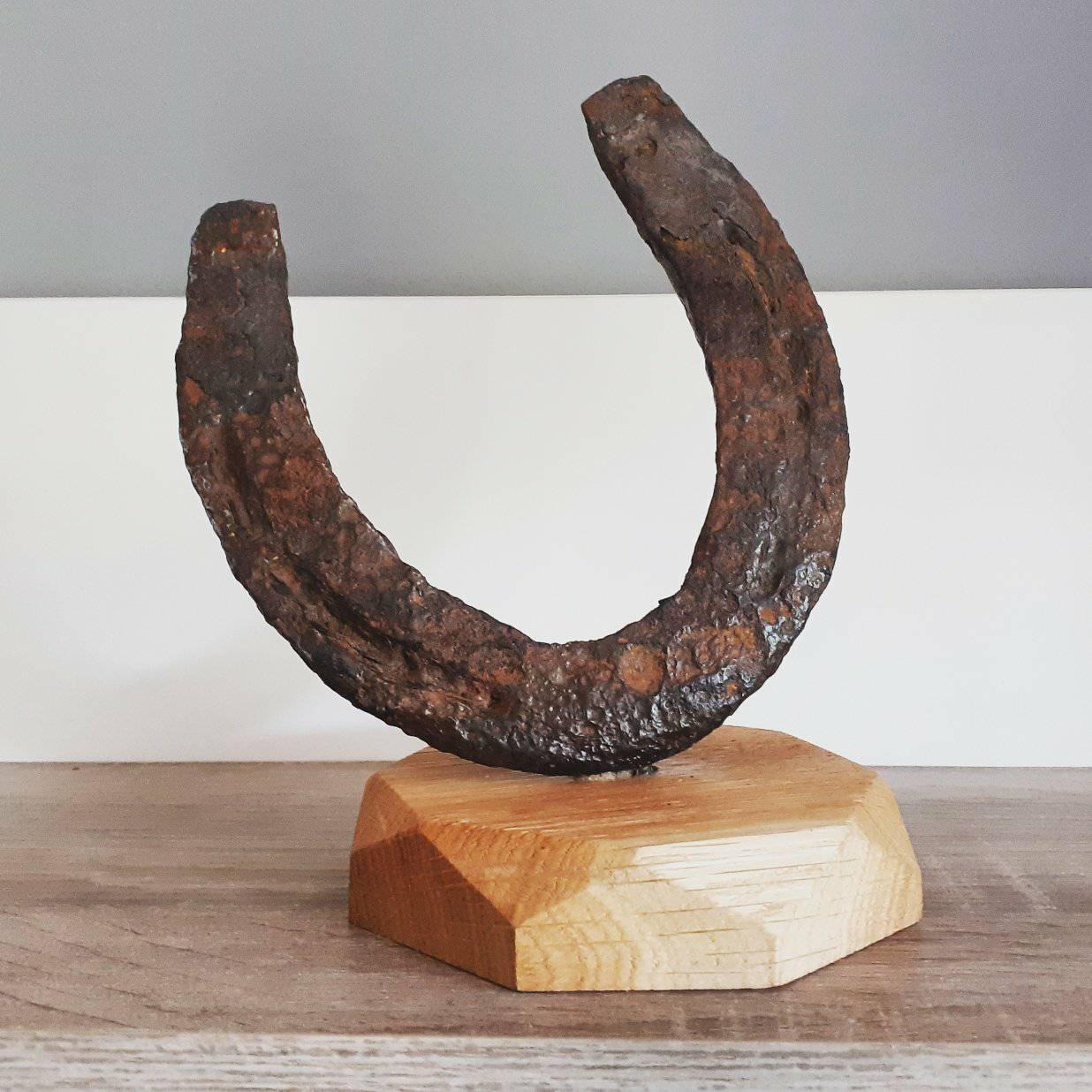 Dekoracyjna, zardzewiała podkowa umieszczona na drewnianej podstawie o ściętych krawędziach, eksponowana na drewnianym blacie.