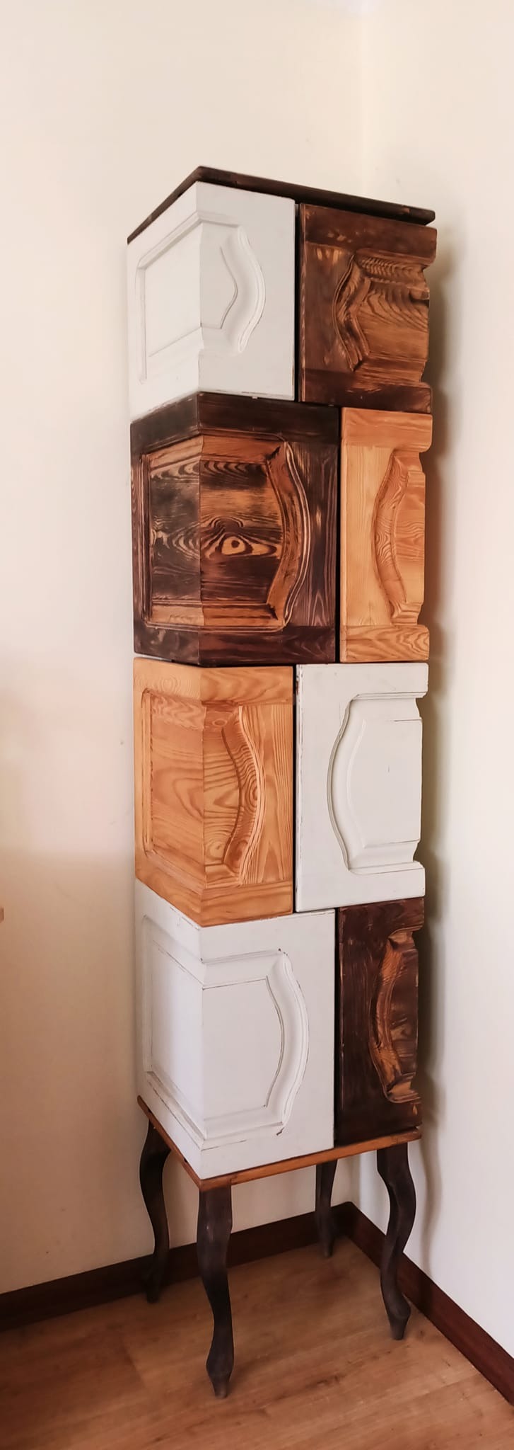 Nietypowa kolumna z drewnianych, kwadratowych modułów z dekoracyjnymi frezami, w kolorach naturalnego drewna i bieli, ustawiona na niskim stoliku z giętymi nogami.