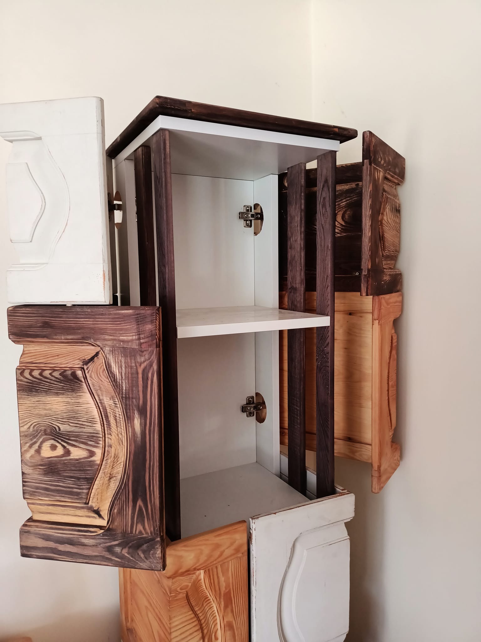 Nietypowa szafka wisząca z otwartymi drzwiczkami, wykonana z różnych rodzajów drewna, z widocznymi zawiasami i półkami wewnątrz.