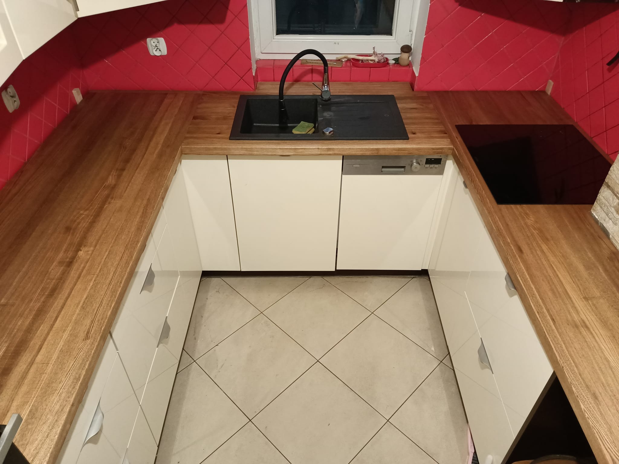 Kątowe ujęcie na nową kuchnię z drewnianym blatem, czarnym zlewem i płytą indukcyjną, białe szafki z szarymi uchwytami, czerwone płytki na ścianie nad blatem.