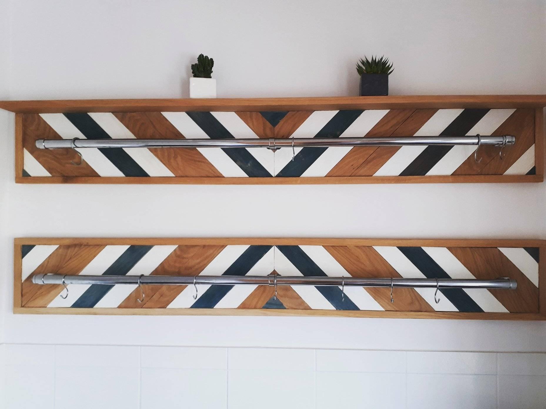 Dwie drewniane półki z geometrycznym wzorem w kolorach drewna, bieli i granatu, z metalowymi rurkami i haczykami, na których można zawieszać ubrania. Na górnej półce stoją małe rośliny w doniczkach.