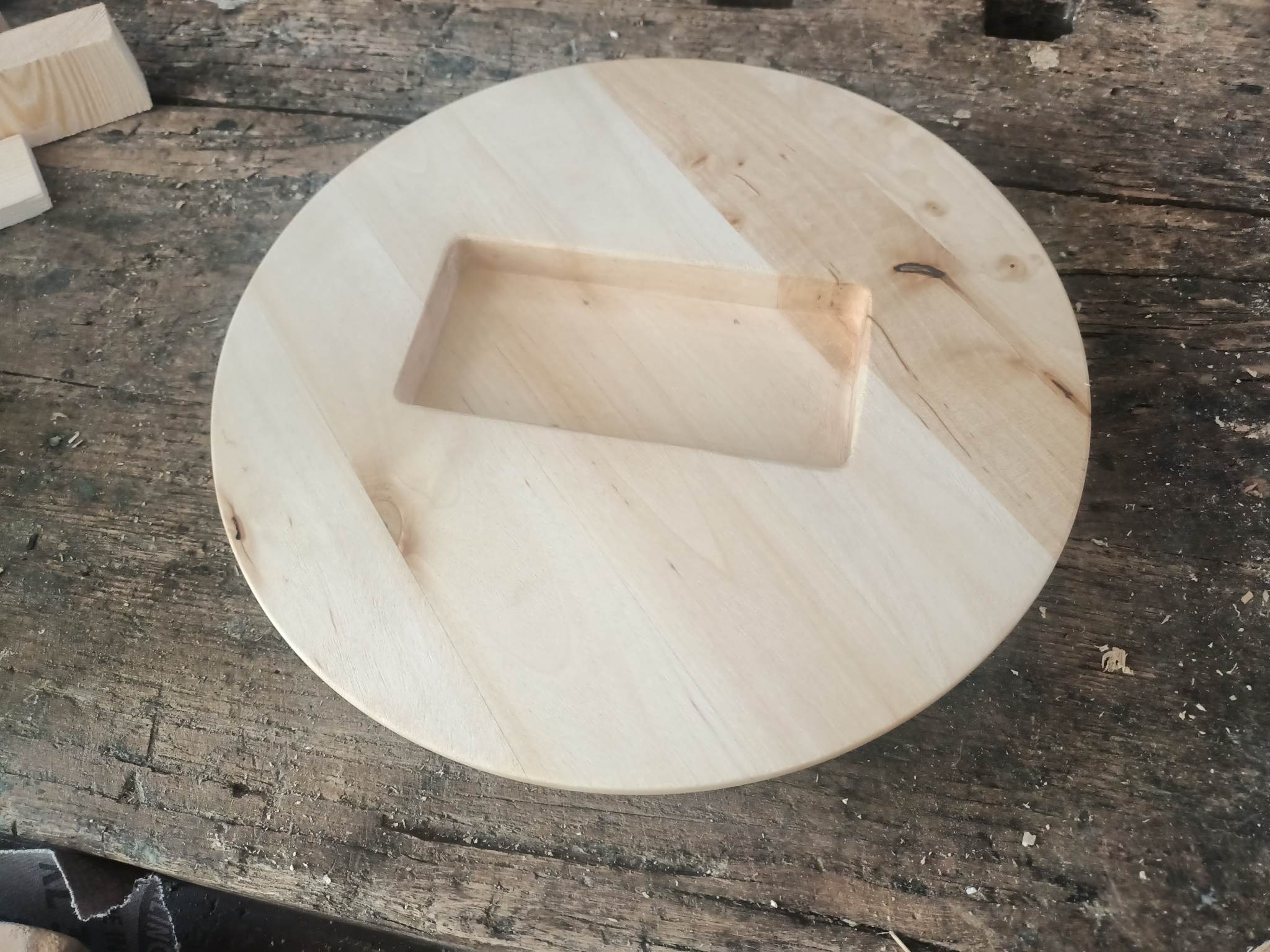 Okrągły, jasny element drewniany z wyfrezowanym prostokątnym wgłębieniem, leżący na roboczym stole z widocznymi śladami obróbki.