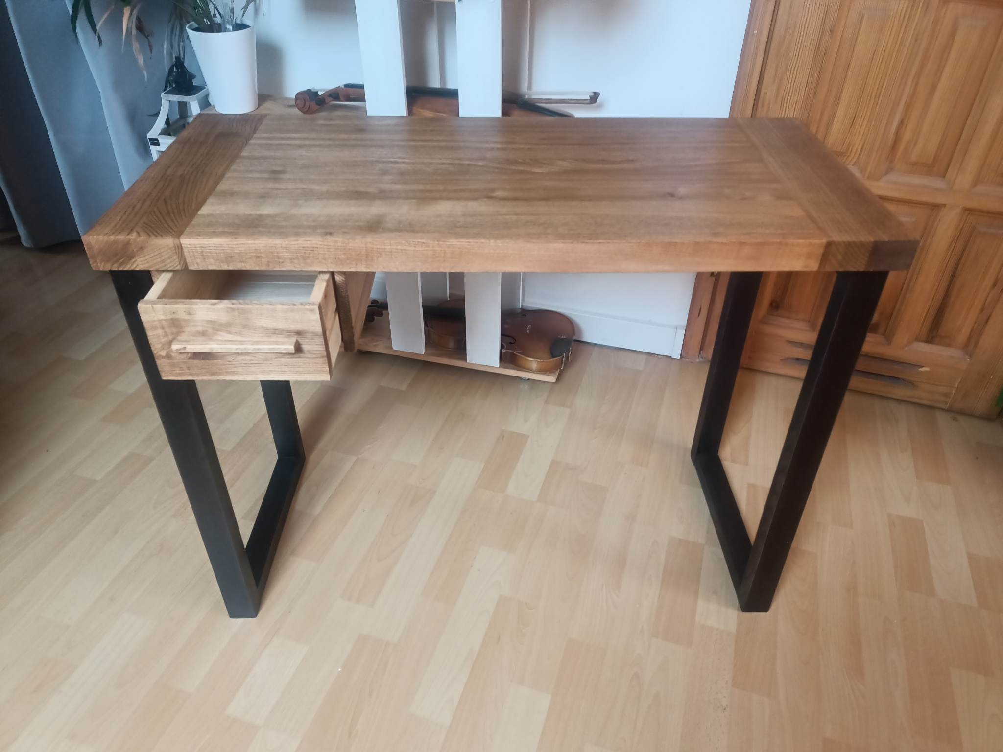 Biurko z blatem z jasnego drewna i czarnymi, metalowymi nogami w kształcie litery V, z wysuwaną szufladą po lewej stronie, na podłodze panele, w tle skrzypce.