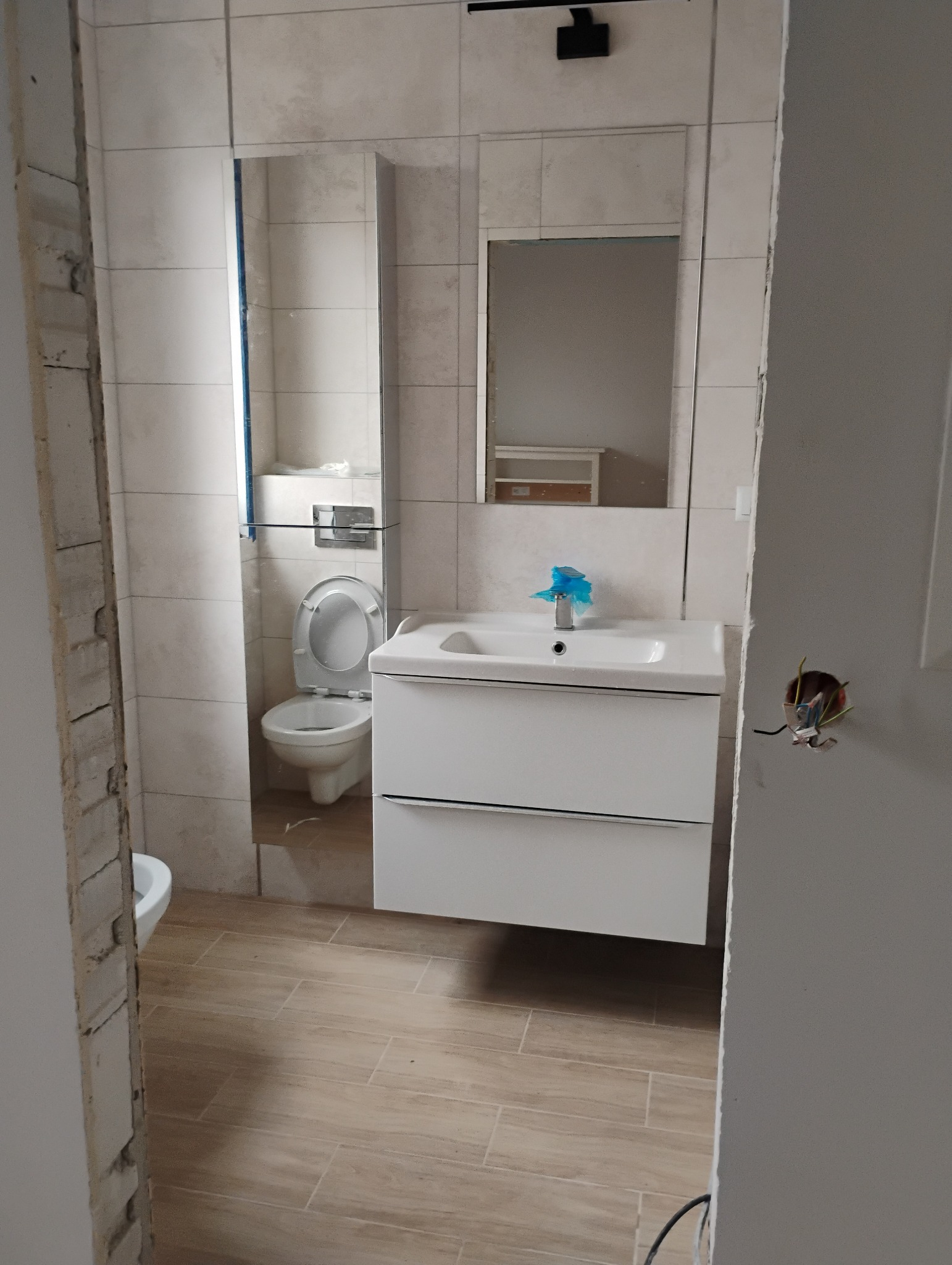 Łazienka w trakcie prac wykończeniowych: widoczna toaleta, umywalka z szafką, lustro oraz niedokończone ściany z wystającymi kablami.