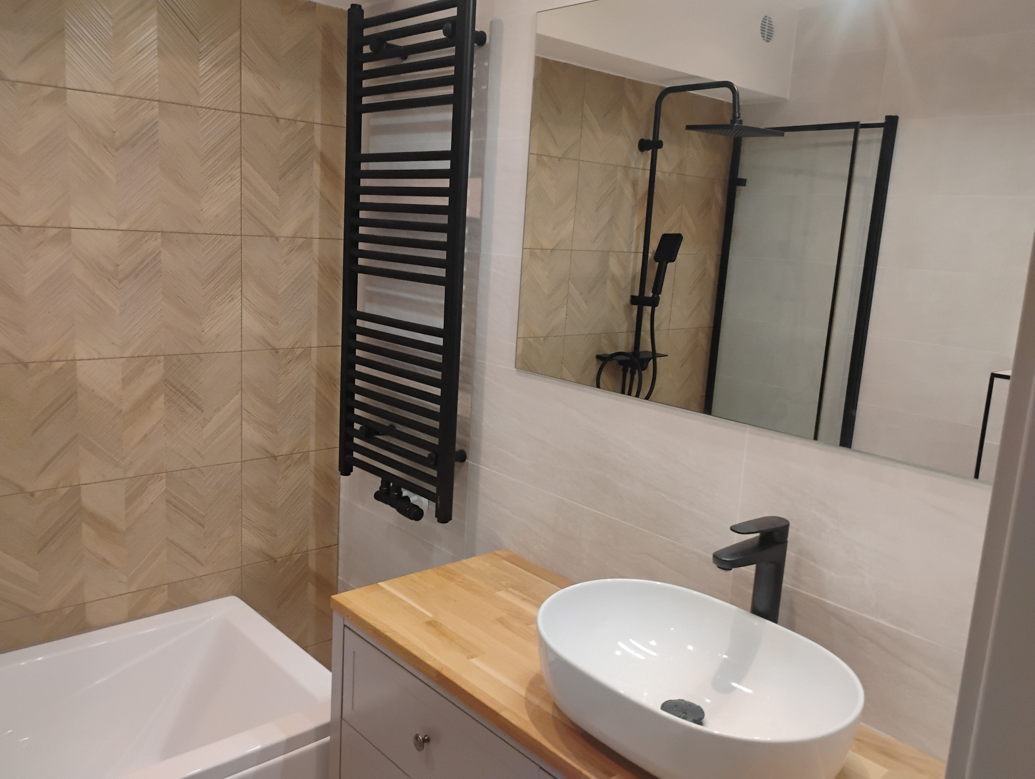 Nowoczesna łazienka z płytkami ułożonymi w jodełkę, czarnym grzejnikiem drabinkowym, umywalką nablatową i czarną armaturą, odbijająca się w lustrze kabina prysznicowa.