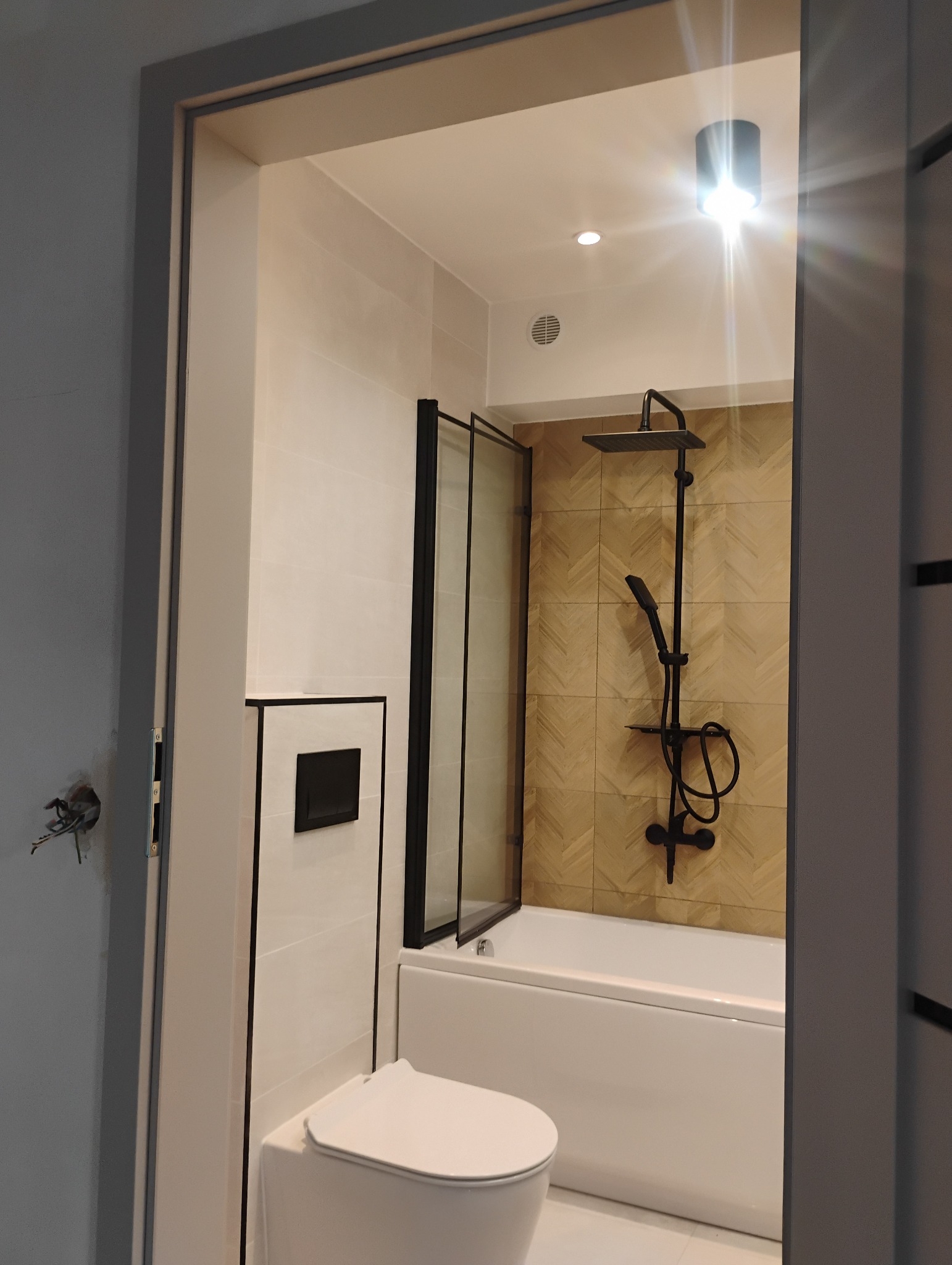 Nowoczesna łazienka z białą ceramiką, czarną armaturą i akcentem drewnopodobnych płytek ułożonych w jodełkę na ścianie w strefie prysznicowej, widok z perspektywy wejścia.