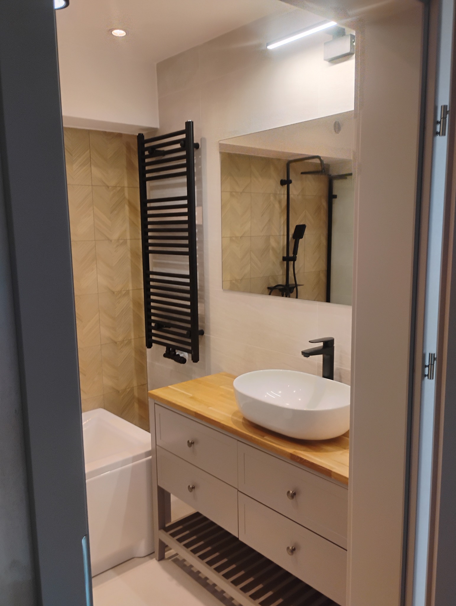 Nowoczesna łazienka z umywalką nablatową, drewnianym blatem, grzejnikiem drabinkowym w kolorze czarnym i płytkami w jodełkę na ścianie, widok przez otwarte drzwi.
