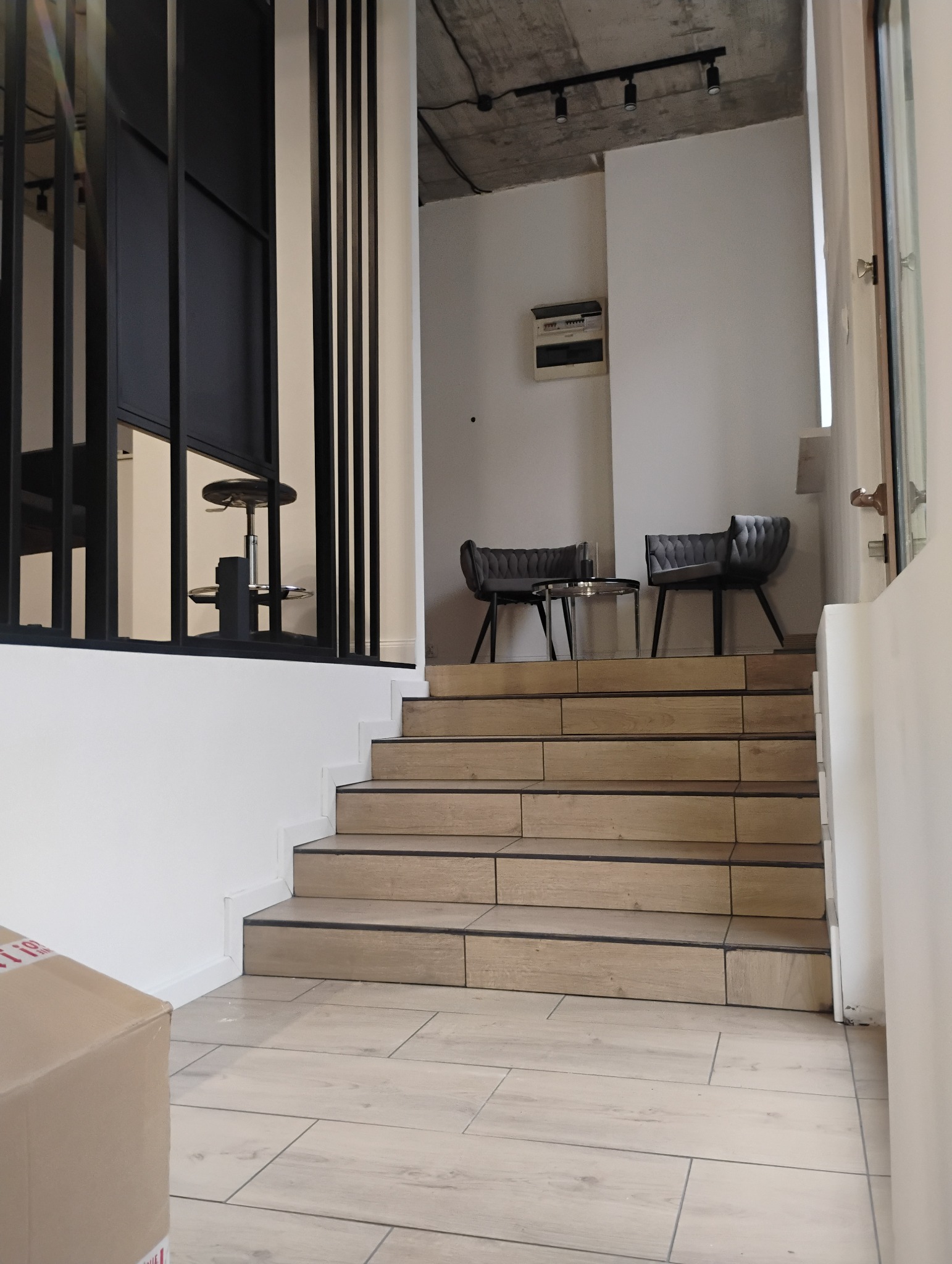Schody wyłożone płytkami imitującymi drewno, prowadzące do strefy relaksu z dwoma szarymi krzesłami i szklanym stolikiem, widok z dołu, częściowo zasłonięty kartonem.