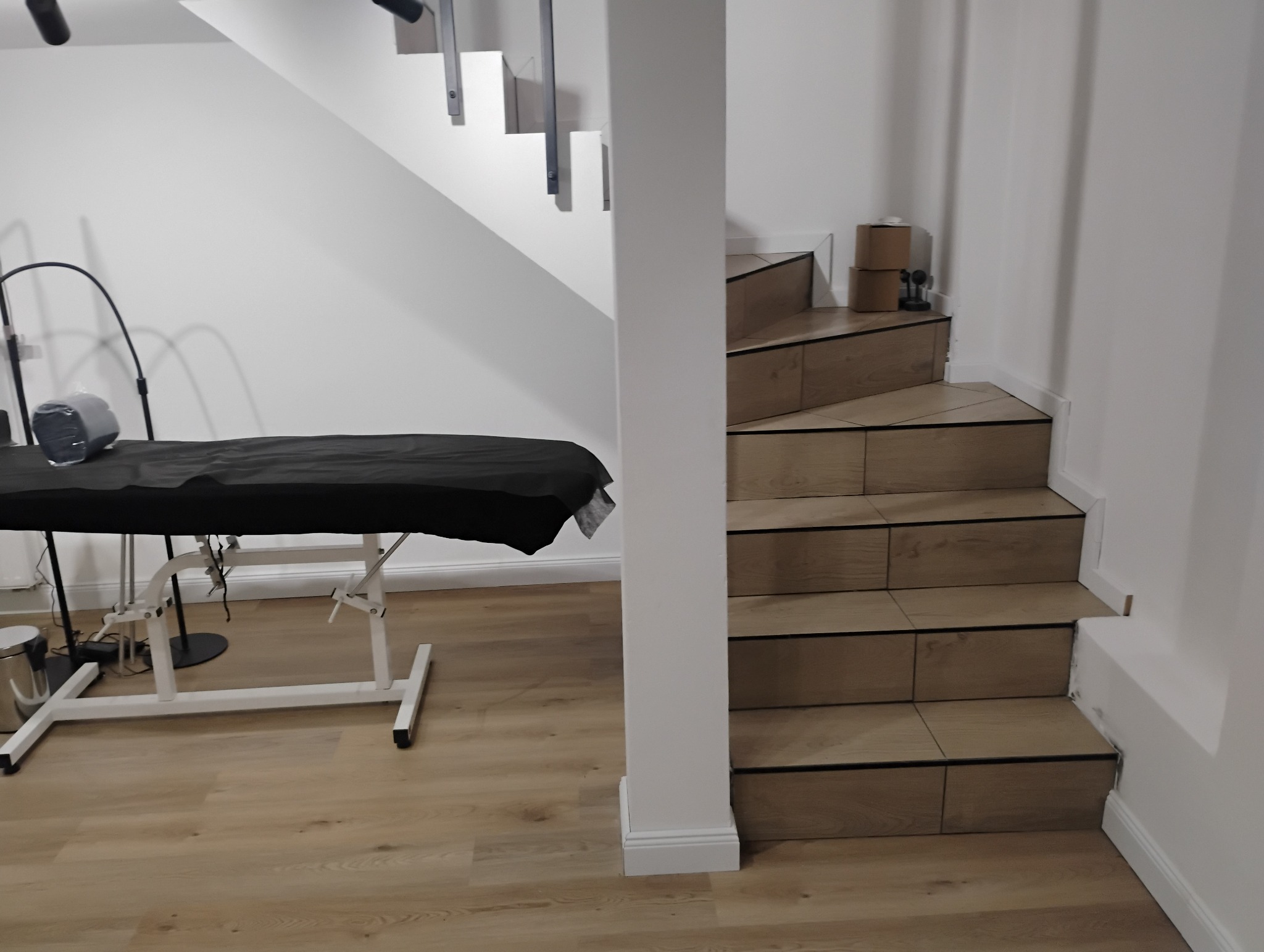 Nowoczesne schody wyłożone panelami imitującymi drewno, z czarnymi akcentami, znajdujące się w jasnym wnętrzu, obok kolumny i zarysu czarnego leżaka kosmetycznego z lampą.