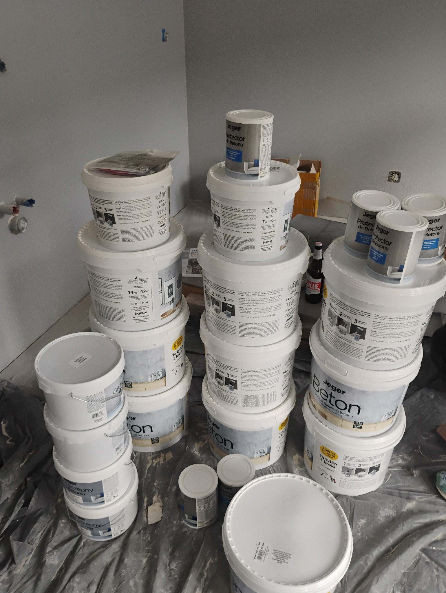 Stos wiaderek z farbą i gruntem marki Jeger na folii malarskiej w trakcie prac wykończeniowych, widoczna butelka piwa.