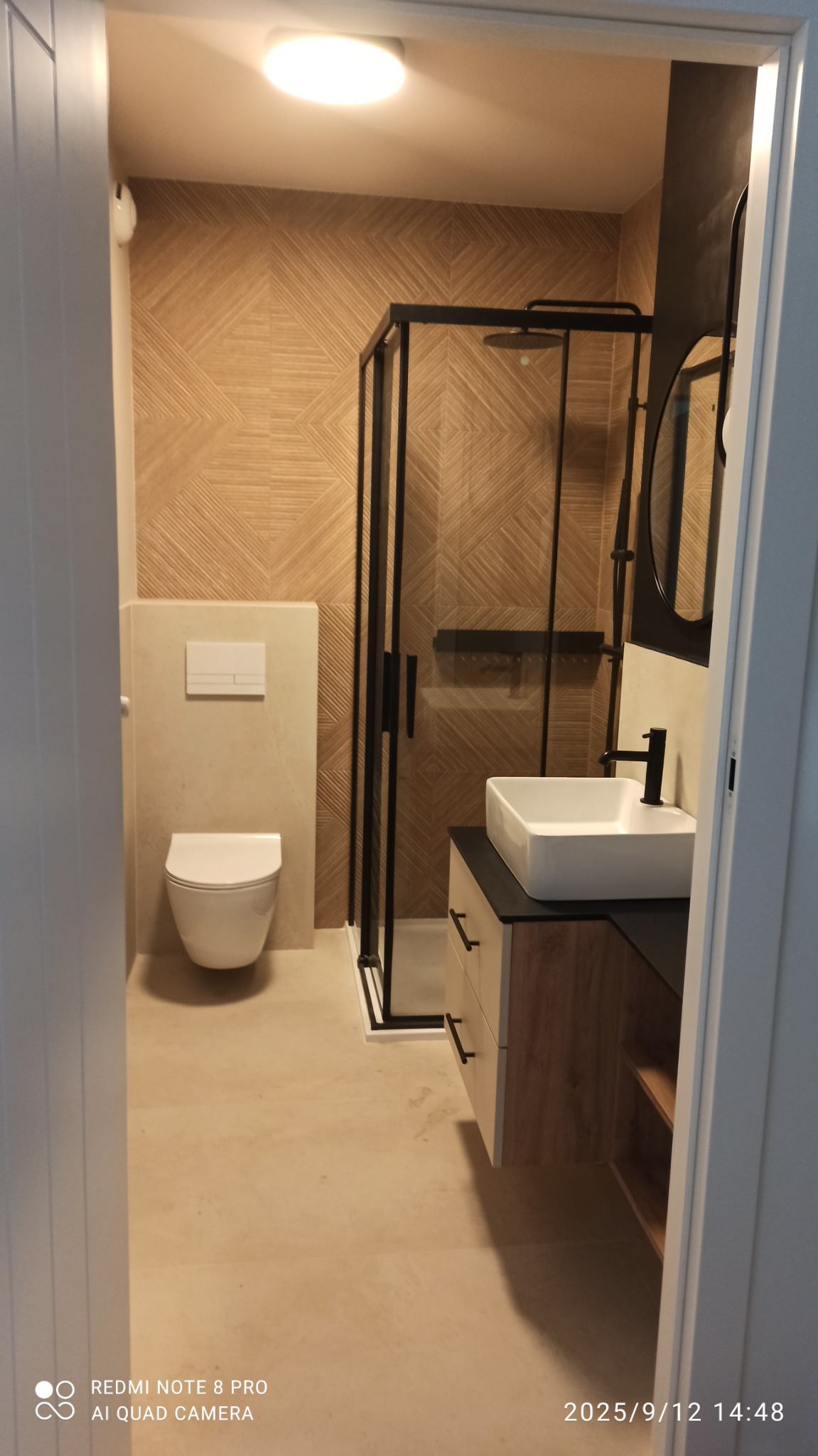 Nowoczesna łazienka z kabiną prysznicową w czarnej ramie, umywalką na szafce z drewnianym frontem i toaletą. Ściany wyłożone płytkami imitującymi drewno.