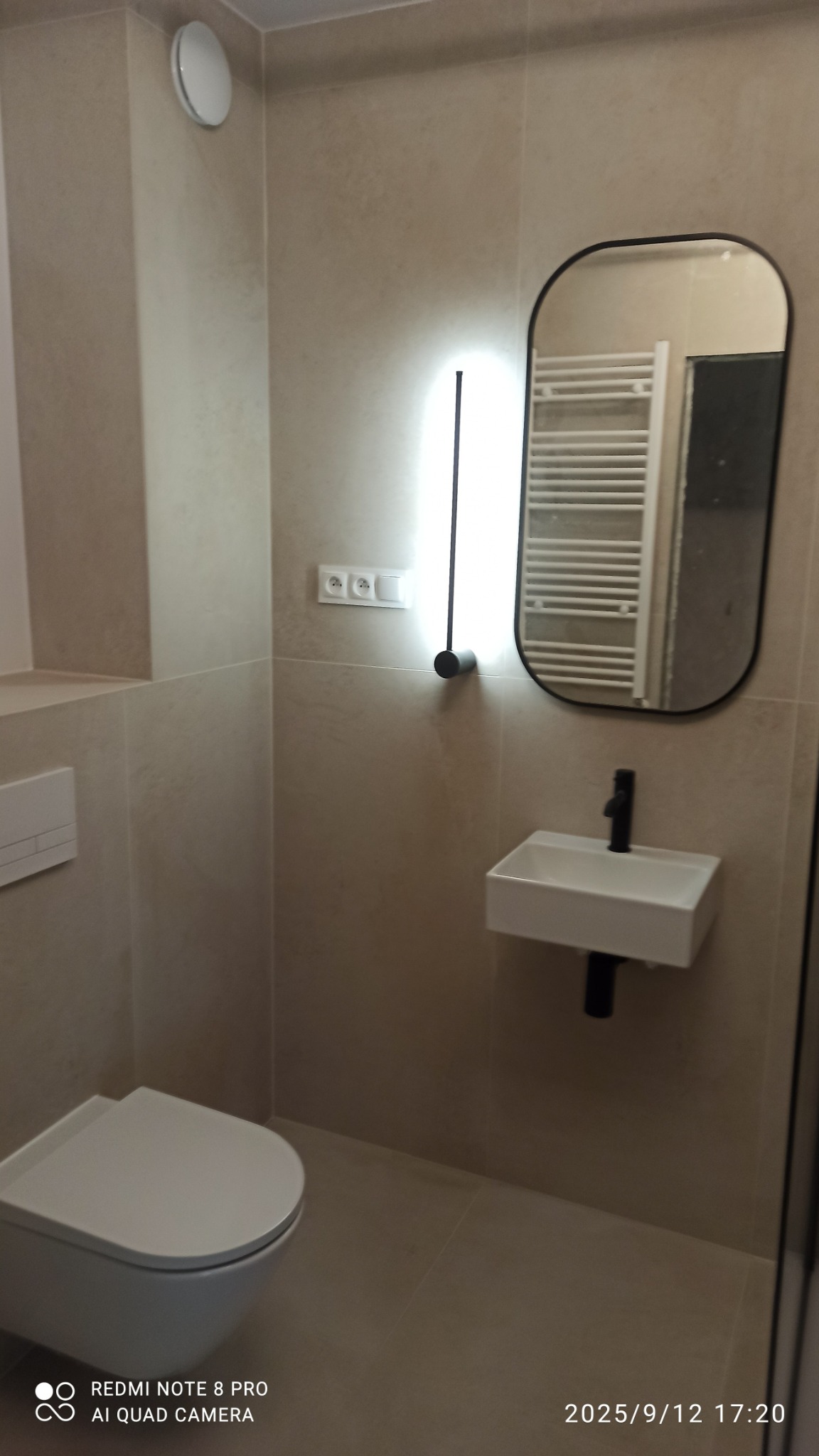 Nowoczesna łazienka w beżowych odcieniach: podwieszana toaleta, mała umywalka z czarną armaturą i podświetlane lustro. Minimalistyczny design z płytkami imitującymi kamień.