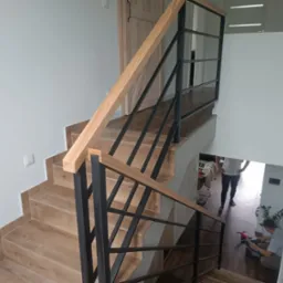 Nowoczesne schody z drewnianymi stopniami i czarną, metalową balustradą, widoczne z g&oacute;ry. W tle osoby na piętrze i roślina doniczkowa.