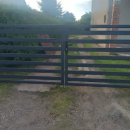 Nowoczesna, pozioma brama wjazdowa w kolorze grafitowym, zamontowana na ceglanych słupkach. Widoczne zarośla w tle, numer domu 41.