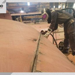 Balustrady, konstrukcje Bydgoszcz i okolice WBO Bartosz Winowiecki - Pracownik w kombinezonie spawalniczym łączy dwie duże, zakrzywione płyty metalowe. Widoczny spaw, sprzęt spawalniczy i fragment hali produkcyjnej.