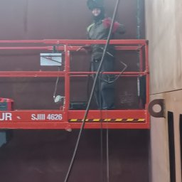 Balustrady, konstrukcje Bydgoszcz i okolice WBO Bartosz Winowiecki - Pracownik w masce spawalniczej na podnośniku, wykonuje prace spawalnicze na dużej, metalowej konstrukcji w hali. Widoczne elementy konstrukcji i narzędzia.