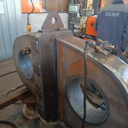 Balustrady, konstrukcje Bydgoszcz i okolice WBO Bartosz Winowiecki - Spawana, metalowa konstrukcja z otworami i uchwytem, widoczna w warsztacie. Ślady spawania i obróbki, spawacz w tle.