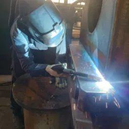 Pracownik w masce spawalniczej łączy metalowy element, iskrząc podczas spawania. Widoczne rękawice ochronne i źródło intensywnego światła.