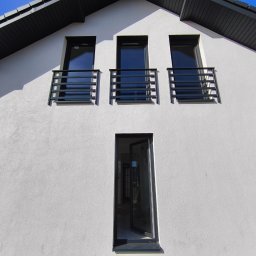 Balustrady, konstrukcje Bydgoszcz i okolice WBO Bartosz Winowiecki - Elewacja domu z trzema balkonami z czarnymi, prostymi balustradami. Otwarty wysoki, wąski otwór drzwiowy. Biały tynk o wyraźnej fakturze. Ciemna podbitka dachowa.