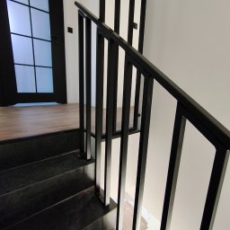 Balustrady, konstrukcje Bydgoszcz i okolice WBO Bartosz Winowiecki - Nowoczesne schody z czarnymi stopniami i minimalistyczną, metalową poręczą. Widok z góry, podkreślający geometrię i kontrast bieli ścian z ciemnym wykończeniem.