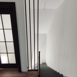 Balustrady, konstrukcje Bydgoszcz i okolice WBO Bartosz Winowiecki - Nowoczesne schody z czarną, minimalistyczną balustradą z pionowych prętów, widoczne z góry. Podłoga drewniana, drzwi z przeszkleniami po lewej stronie.