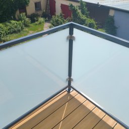 Balustrady, konstrukcje Bydgoszcz i okolice WBO Bartosz Winowiecki - Narożnik nowoczesnej balustrady balkonowej z matowego szkła i ciemnych metalowych profili, zamontowanej na drewnianym tarasie kompozytowym, z widokiem na zielony ogród.