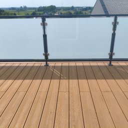 Balustrady, konstrukcje Bydgoszcz i okolice WBO Bartosz Winowiecki - Jasnobrązowa deska kompozytowa na balkonie z nowoczesną balustradą z ciemnego metalu i matowego szkła, w słoneczny dzień z widokiem na zieleń.