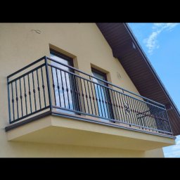 Balustrady, konstrukcje Bydgoszcz i okolice WBO Bartosz Winowiecki - Czarna metalowa balustrada balkonowa z ozdobnymi kulkami na pionowych prętach i centralnym medalionem, zamontowana na jasnobeżowej elewacji budynku pod błękitnym niebem.