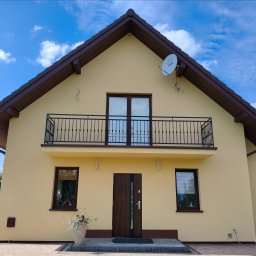 Balustrady, konstrukcje Bydgoszcz i okolice WBO Bartosz Winowiecki - Jasnożółta fasada nowoczesnego domu z brązowym dachem i oknami. Na piętrze balkon z czarną, metalową balustradą z ozdobnymi tralkami i centralnym motywem. Przed wejściem donica z kwiatami...
